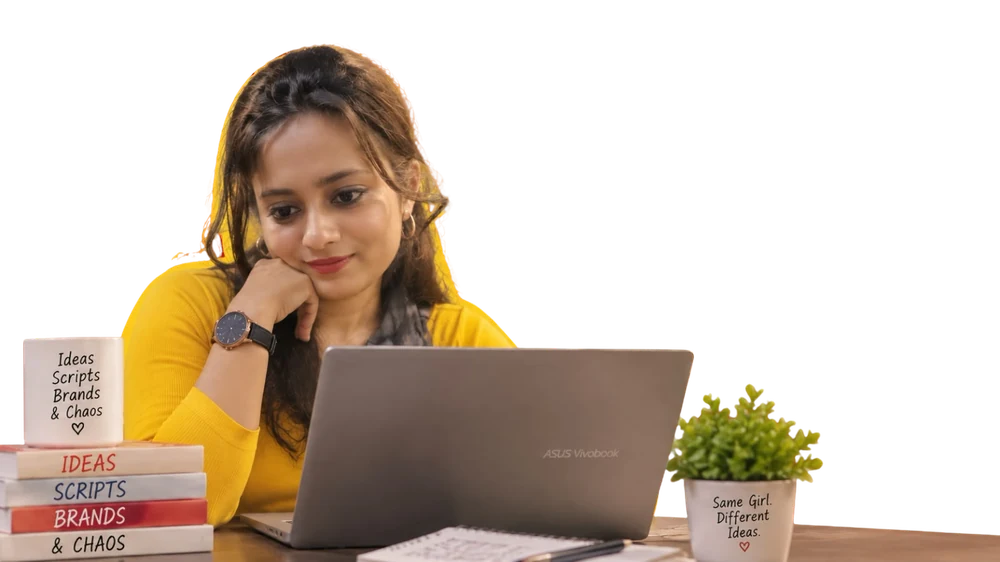 Kausshiki at her desk in a yellow top, resting her chin on her hand and looking at a laptop, next to a mug and a stack of books reading Ideas, Scripts, Brands and Chaos